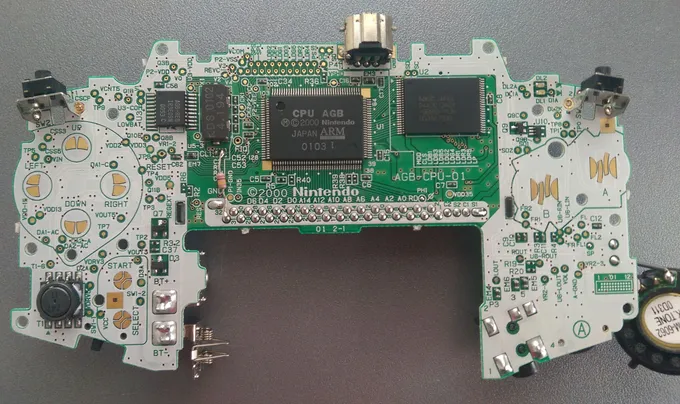 Vista frontal da placa-mãe do Game Boy Advance, com a CPU AGB ao centro, o chip de memória à direita e os contatos dos botões nas laterais