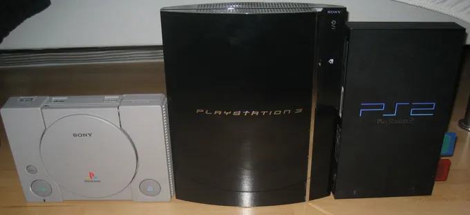 Consoles PlayStation 1, PlayStation 3 e PlayStation 2 lado a lado sobre uma mesa, da esquerda para a direita