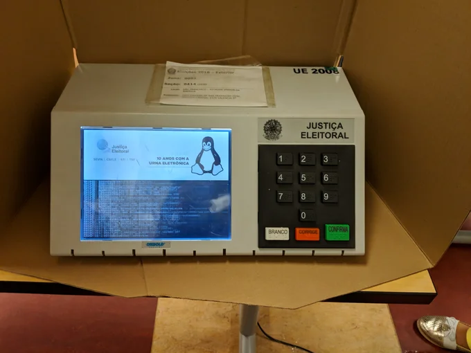 Urna eletrônica brasileira durante a inicialização, com mensagens do sistema na tela à esquerda e o teclado à direita